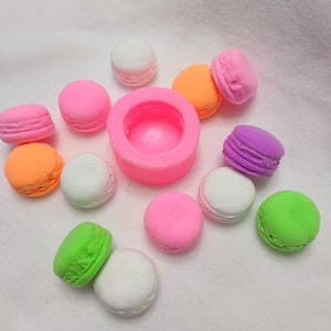 May include: A pink silicone mold for making macarons, surrounded by colorful macarons in various shades of pink, green, white, and orange.
