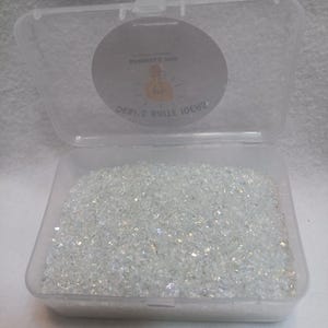 May include: A clear plastic container filled with iridescent, crushed glass pieces. The container is open, revealing the contents. The lid has a circular sticker with text and a lightbulb graphic. The text on the sticker reads "SHIMMER MIX" and "DEBI.2 BRILE IDEAS".