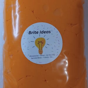 May include: A clear plastic bag filled with bright orange air-dry clay. A circular label displays the text "Brite Ideas" with a lightbulb illustration and lists craft supplies like rhinestones and glitter.