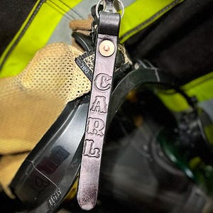 May include: A dark leather keychain with the name "CARL" embossed in capital letters. The keychain is attached to a silver clasp and a metal chain. The background includes a firefighter's gear, with yellow and black reflective stripes.