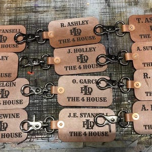 May include: A collection of personalized leather keychains. Each keychain is rectangular, with rounded corners, and features a name and the text "THE 4 HOUSE" in black. The keychains are connected with metal clasps and leather straps.
