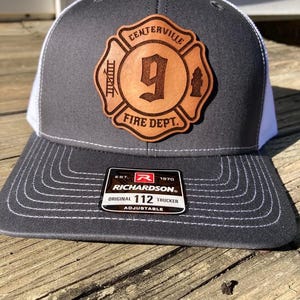 May include: A gray and white trucker hat with a brown leather patch that says "Centerville Fire Dept. 9". The hat is made by Richardson and is adjustable.