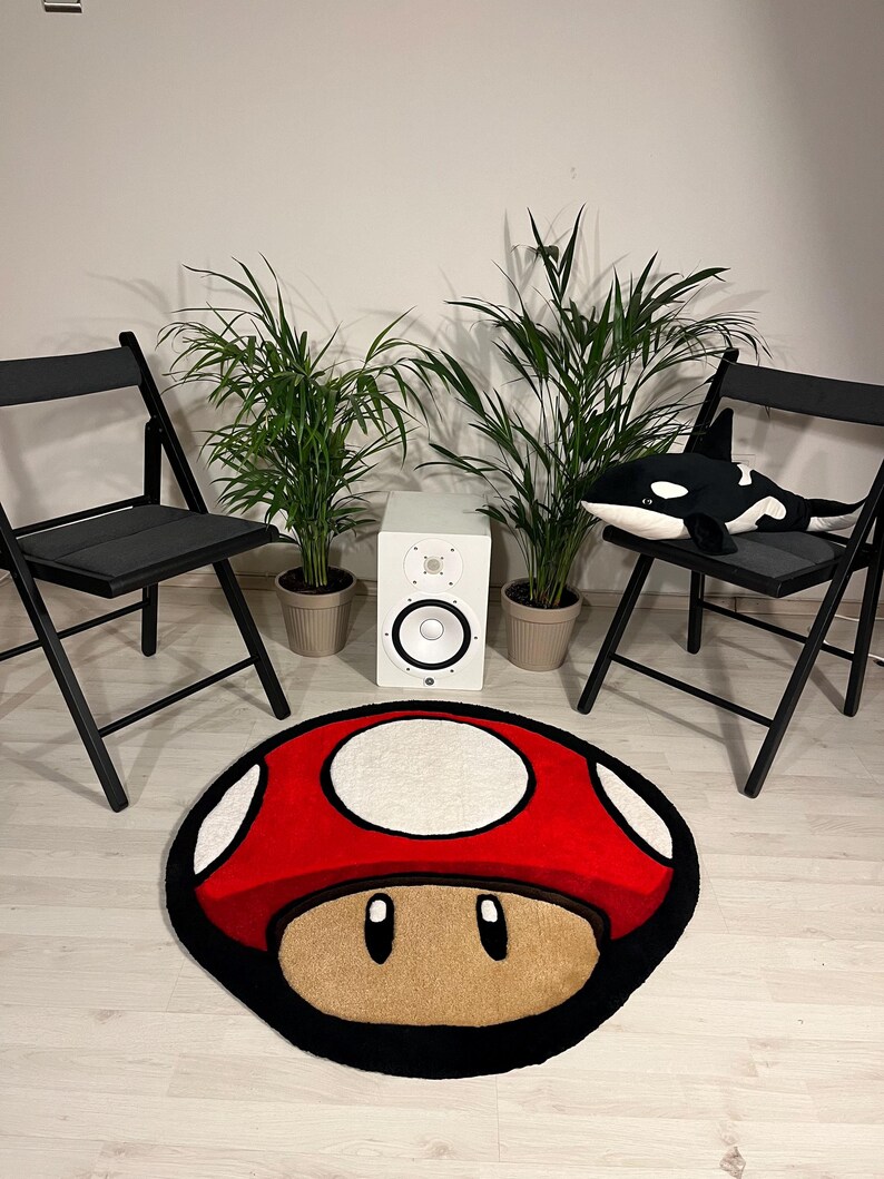 May include: A red and white mushroom-shaped rug with a black outline. The rug is on a light wood floor. There are two black folding chairs and two potted plants in the background.