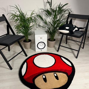 May include: A red and white mushroom-shaped rug with a black outline. The rug is on a light wood floor. There are two black folding chairs and two potted plants in the background.