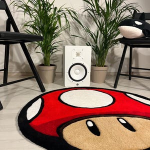 May include: A red and white mushroom-shaped rug with black trim. The rug is on a wooden floor in front of a white wall. There are two potted plants and two black folding chairs in the background.