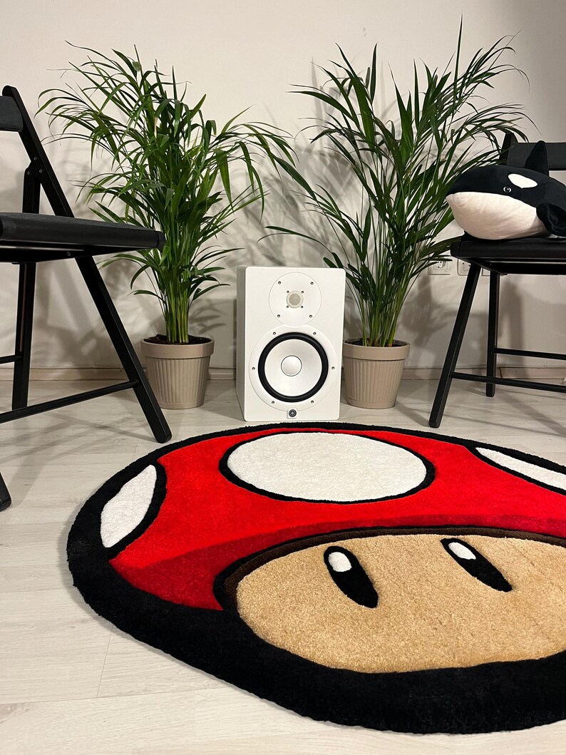 May include: A red and white mushroom-shaped rug with black trim. The rug is on a wooden floor in front of a white wall. There are two potted plants and two black folding chairs in the background.
