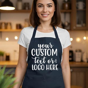 May include: A navy blue apron with the text "your CUSTOM TEXT or LOGO HERE" in white script. The apron has a bib and a pocket. The person in the image is wearing a white t-shirt.