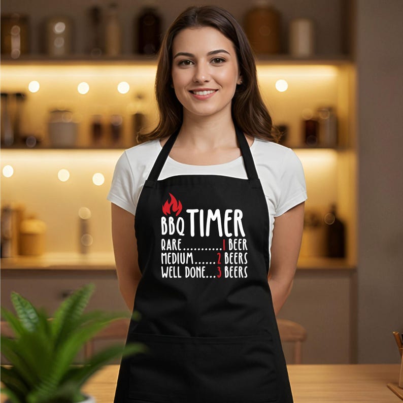 May include: Black apron with white text and a red flame graphic. The text reads "BBQ TIMER" with beer recommendations for rare, medium, and well-done. The apron has a pocket and black neck and waist straps.