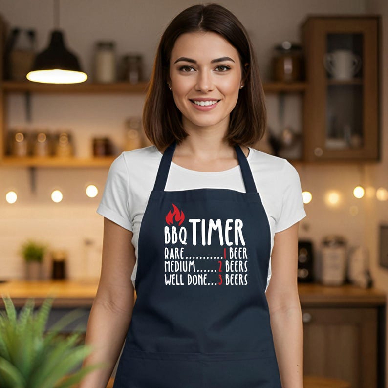 May include: A navy blue apron with the text "BBQ TIMER" and a flame graphic. The text below lists "Rare" with 1 beer, "Medium" with 2 beers, and "Well Done" with 3 beers. The apron is worn over a white t-shirt.