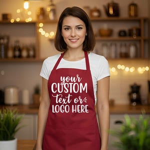 May include: A burgundy apron with white text that reads "your CUSTOM Text or LOGO HERE". The apron is worn over a white t-shirt. The background is a kitchen with wooden shelves and warm lighting.