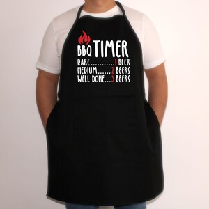 May include: Black apron with the text "BBQ TIMER" and a flame graphic. The text below reads "Rare...1 Beer, Medium...2 Beers, Well Done...3 Beers". The apron has two front pockets and a neck strap.