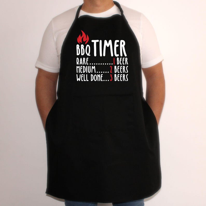 May include: Black apron with the text "BBQ TIMER" and a flame graphic. The text below reads "Rare...1 Beer, Medium...2 Beers, Well Done...3 Beers". The apron has two front pockets and a neck strap.