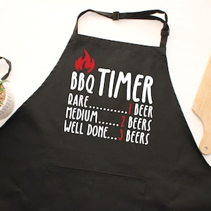 May include: Black BBQ apron with the text "BBQ TIMER" and a flame graphic. The text below indicates beer consumption levels: Rare - 1 beer, Medium - 2 beers, Well Done - 3 beers. Includes a pocket, a small plant, and a wooden cutting board.