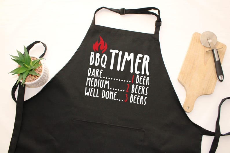 May include: Black BBQ apron with the text "BBQ TIMER" and a flame graphic. The text below indicates beer consumption levels: Rare - 1 beer, Medium - 2 beers, Well Done - 3 beers. Includes a pocket, a small plant, and a wooden cutting board.