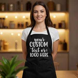 May include: Black apron with the text "your CUSTOM Text or LOGO HERE" in white script. The apron has a pocket and black straps. The person is wearing a white t-shirt.