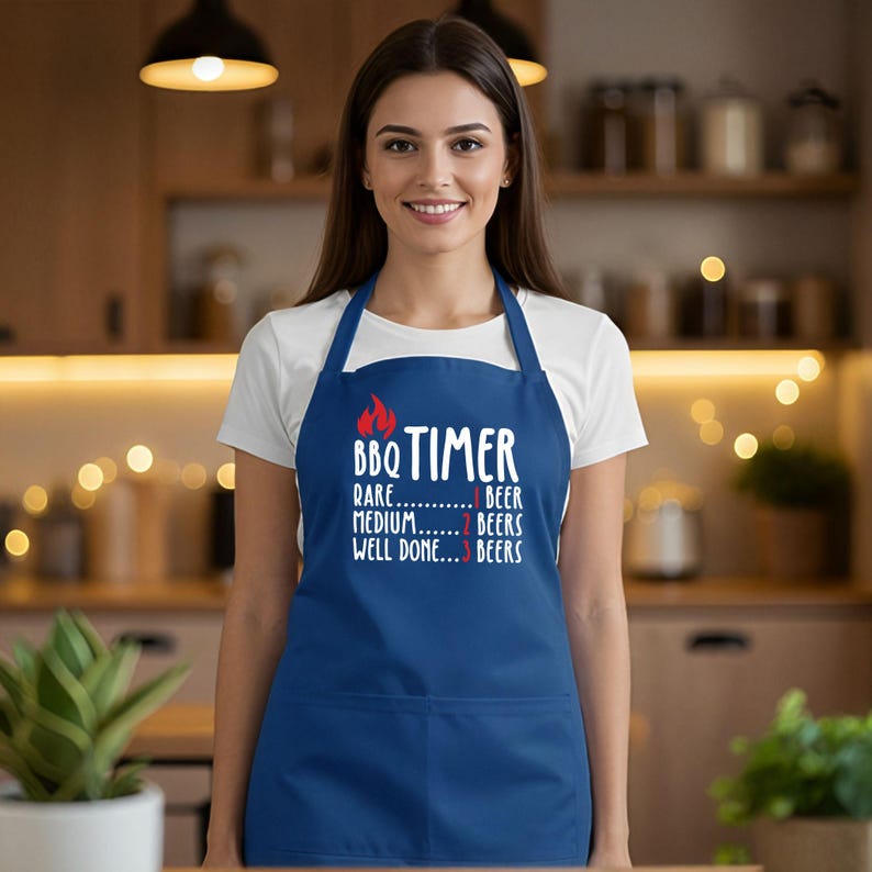 May include: A navy blue apron with the text "BBQ TIMER" and a flame graphic. The text below indicates beer consumption levels: Rare - 1 beer, Medium - 2 beers, Well Done - 3 beers. The apron has a front pocket and neck and waist straps.