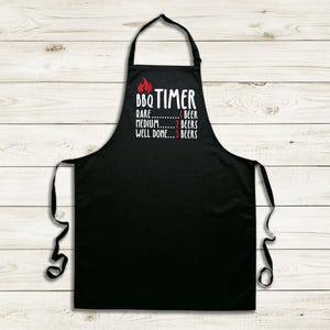 May include: Black apron with white text and a red flame graphic. The text reads "BBQ TIMER" above a cooking guide: "Rare...1 Beer, Medium...2 Beers, Well Done...3 Beers". The apron has a neck loop and waist ties.
