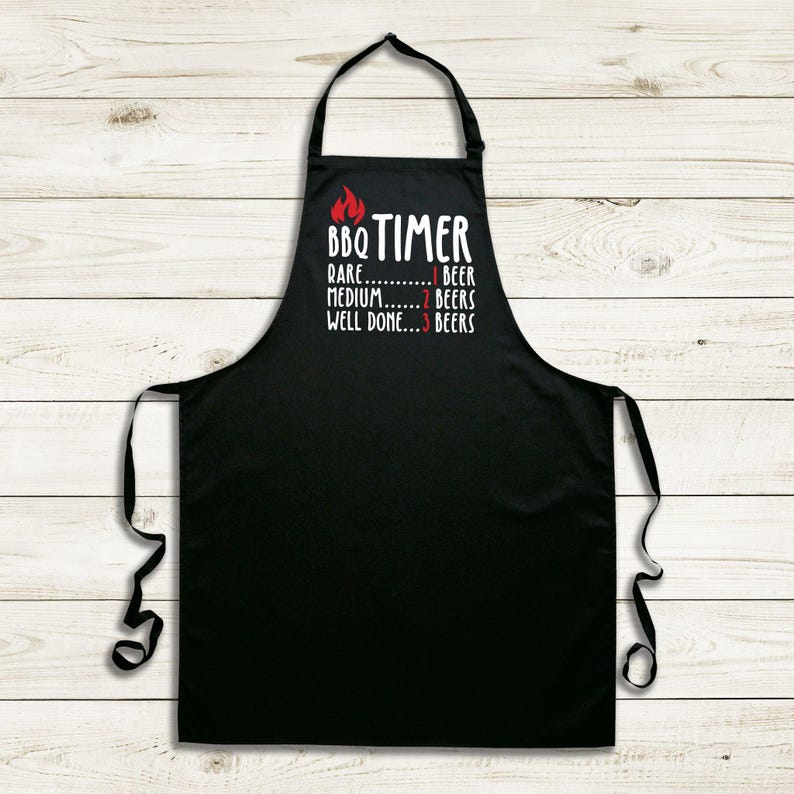 May include: Black apron with white text and a red flame graphic. The text reads "BBQ TIMER" above a cooking guide: "Rare...1 Beer, Medium...2 Beers, Well Done...3 Beers". The apron has a neck loop and waist ties.