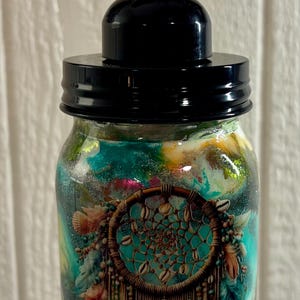 May include: A decorative soap dispenser with a black pump and lid. The glass jar features a dreamcatcher design with turquoise, brown, and gold accents. The jar is filled with a colorful, swirling liquid.
