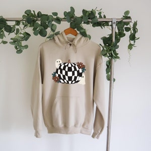 May include: A beige hoodie with a checkered pumpkin and ghosts graphic. The pumpkin is black and white with a brown stem. The ghosts are white with black eyes. There are brown and blue flowers around the pumpkin.