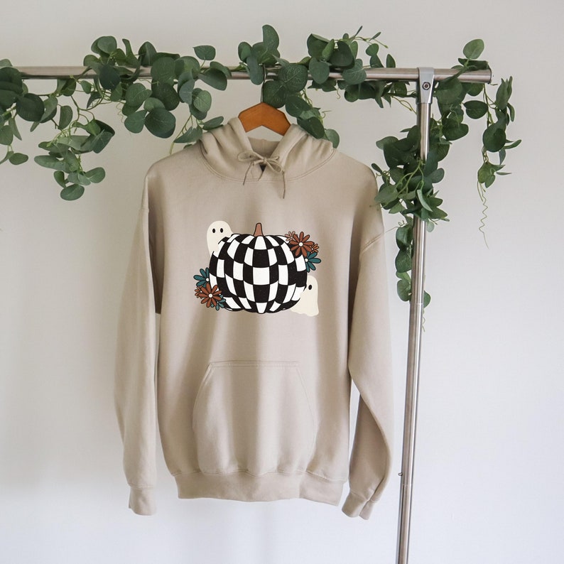 May include: A beige hoodie with a checkered pumpkin and ghosts graphic. The pumpkin is black and white with a brown stem. The ghosts are white with black eyes. There are brown and blue flowers around the pumpkin.