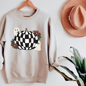 May include: A light gray sweatshirt with a black and white checkered pumpkin design. The pumpkin is surrounded by small brown, blue, and white flowers and two white ghosts.