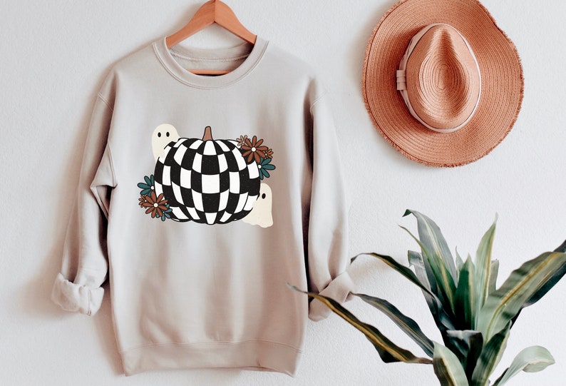 May include: A light gray sweatshirt with a black and white checkered pumpkin design. The pumpkin is surrounded by small brown, blue, and white flowers and two white ghosts.