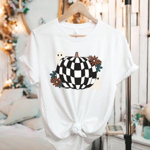 May include: White t-shirt with a black and white checkered pumpkin design. The pumpkin is surrounded by brown, blue, and white flowers and two white ghosts.