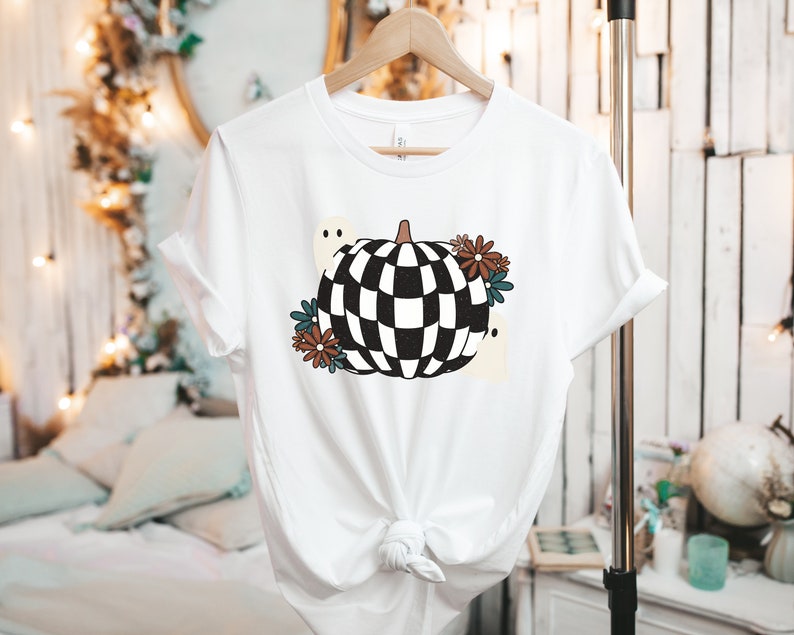 May include: White t-shirt with a black and white checkered pumpkin design. The pumpkin is surrounded by brown, blue, and white flowers and two white ghosts.