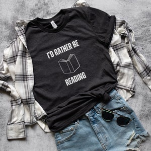 May include: A dark gray t-shirt with the text "I'd rather be reading" and a white outline of a book. The shirt is worn with a white and black plaid shirt and blue denim shorts.