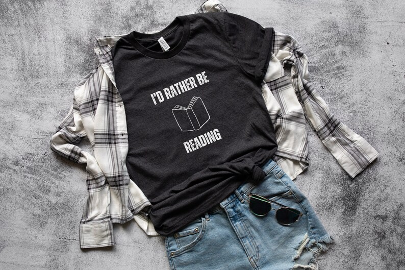 May include: A dark gray t-shirt with the text "I'd rather be reading" and a white outline of a book. The shirt is worn with a white and black plaid shirt and blue denim shorts.