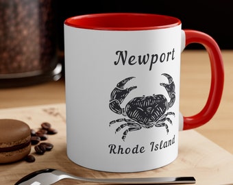 May include: White ceramic mug with a red rim and handle. The mug has a black and white illustration of a crab and the text "Newport Rhode Island".