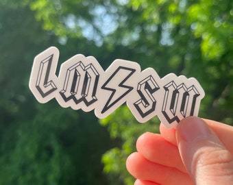 May include: A white sticker with the text "LM⚡️SW" in black, with a lightning bolt between the letters. The sticker is held in a hand against a blurred green background.