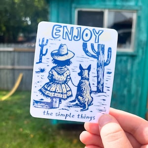May include: A white sticker with blue text and illustrations. The word "ENJOY" is at the top. The image depicts a girl in a dress and hat with a dog, cacti, and the words "the simple things".