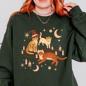 May include: Dark green sweatshirt featuring a whimsical illustration of three cats. One cat wears a witch hat, another a crown, and the third a floral wreath. The design includes candles, moons, and stars, creating a magical theme.