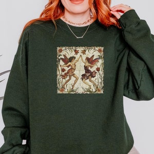May include: A dark green sweatshirt with a vintage illustration of two fairies dancing in a floral frame. The fairies are wearing colorful dresses and have wings.