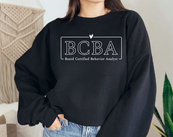 Puede incluir: Una sudadera negra de cuello redondo con texto blanco que dice BCBA y Board Certified Behavior Analyst dentro de un marco rectangular, con un pequeño icono de corazón centrado sobre el texto. La sudadera tiene mangas largas y corte holgado.