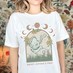 May include: White t-shirt featuring a whimsical illustration of a frog playing a stringed instrument, surrounded by sunflowers and mushrooms. The design includes moon phases and the text "HAPPY JOYOUS & FREE".