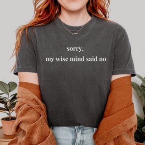 May include: A woman wearing a dark gray t-shirt with the text "sorry, my wise mind said no" printed on it. She is wearing a brown jacket and blue jeans.
