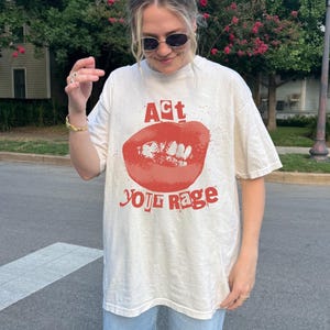 Puede incluir: Una camiseta blanca con un gráfico rojo de una boca con el texto "Act Your Rage" impreso en ella.
