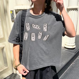 Puede incluir: Una camiseta gris oscuro de manga corta con la frase "GOOD LUCK" escrita con fichas de dominó. La camiseta es ligeramente holgada y tiene un aspecto vintage descolorido. La persona lleva una falda negra y un collar dorado.