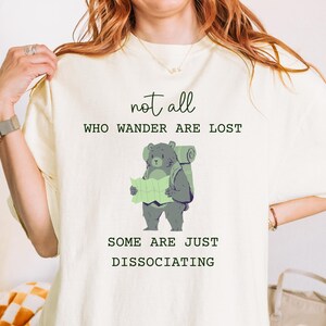 以下が含まれることがあります： 地図を持ったリュックサックを背負ったクマの漫画が描かれた白いTシャツ。シャツのテキストは「not all who wander are lost some are just dissociating.」です。