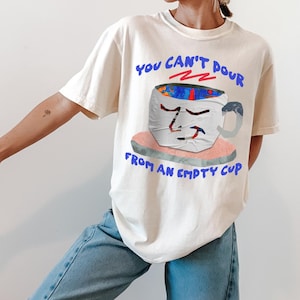 Puede incluir: Camiseta color crema con un gráfico de una taza de café y el texto "YOU CAN'T POUR FROM AN EMPTY CUP" en azul. El diseño de la taza presenta una cara y detalles coloridos. La camiseta se combina con vaqueros azules y zapatillas blancas.
