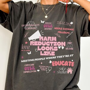 May include: A dark grey t-shirt with a pink graphic print that says "Harm Reduction Looks Like" and includes phrases like "Empathy Over Judgement", "Compassion Over Criminalization", and "Meeting People Where They're At".