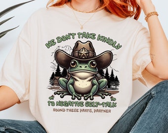 Op de afbeelding: Een beige t-shirt met een groene kikker die een sheriffhoed draagt. Het shirt heeft de tekst "We don't take kindly to negative self-talk. Round these parts, partner."