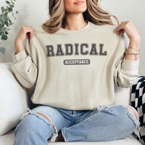 Puede incluir: Una sudadera beige con el texto "RADICAL ACCEPTANCE" en letras negras.