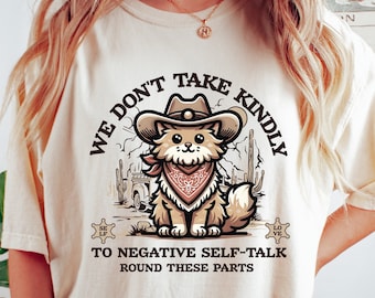 May include: A cream colored t-shirt with a cartoon cat wearing a cowboy hat and bandana. The text "WE DON'T TAKE KINDLY TO NEGATIVE SELF-TALK ROUND THESE PARTS" is printed on the shirt.