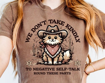 May include: Brown t-shirt with a cartoon cat wearing a cowboy hat and bandana. The text on the shirt reads "We don't take kindly to negative self-talk round these parts".