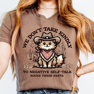 Puede incluir: Camiseta marrón con un gato de dibujos animados que lleva un sombrero de vaquero y un pañuelo. El texto de la camiseta dice "We don't take kindly to negative self-talk round these parts".