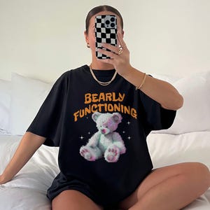 Puede incluir: Camiseta negra de gran tamaño con un gráfico de un oso de peluche blanco y el texto "Bearly Functioning" en naranja.